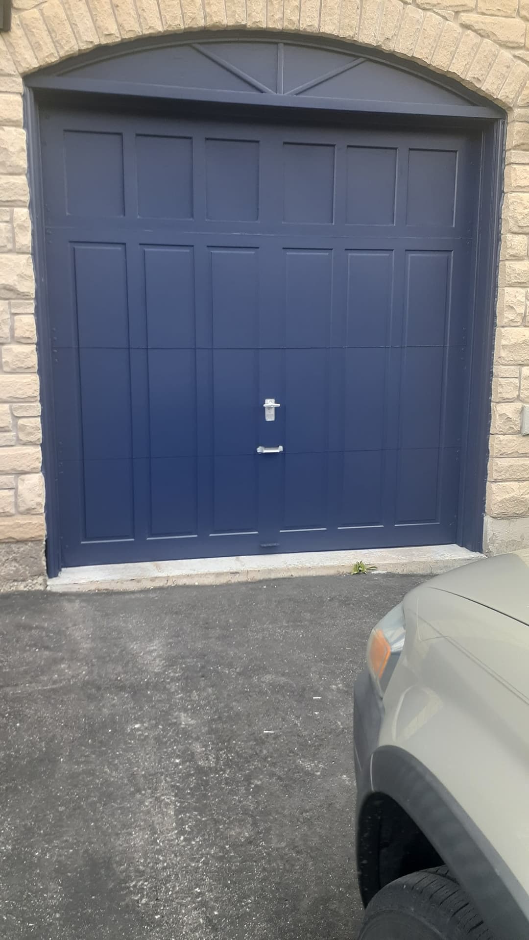 Exterior view of a dark navy-blue paneled garage door set in a light stone facade with an arched top surround. A vehicle is partially visible in the driveway on the right.