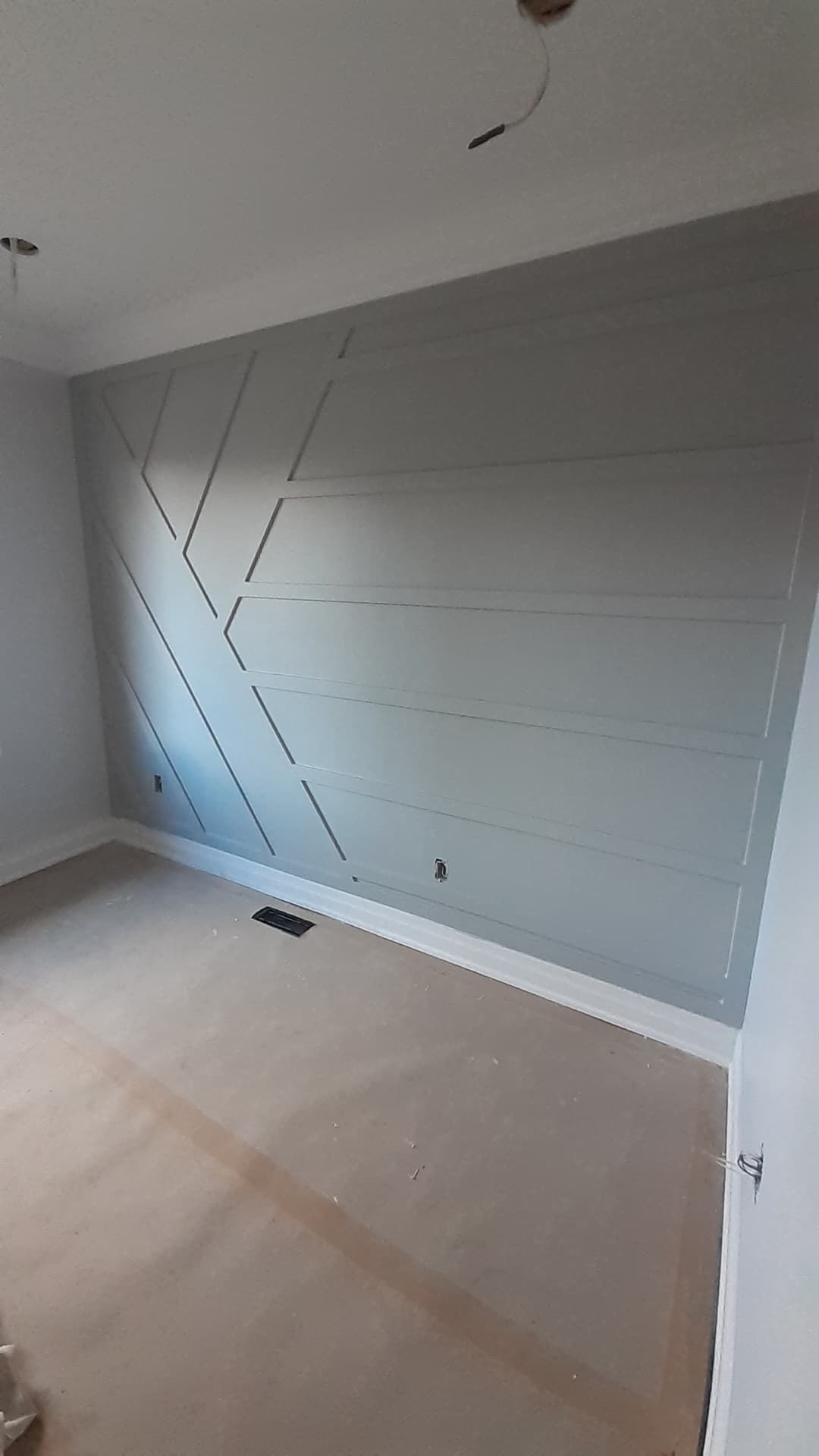 Interior room with a gray feature wall made of long horizontal and angled trim pieces forming a modern geometric pattern. The floor is covered for construction, with unfinished ceiling light openings visible.