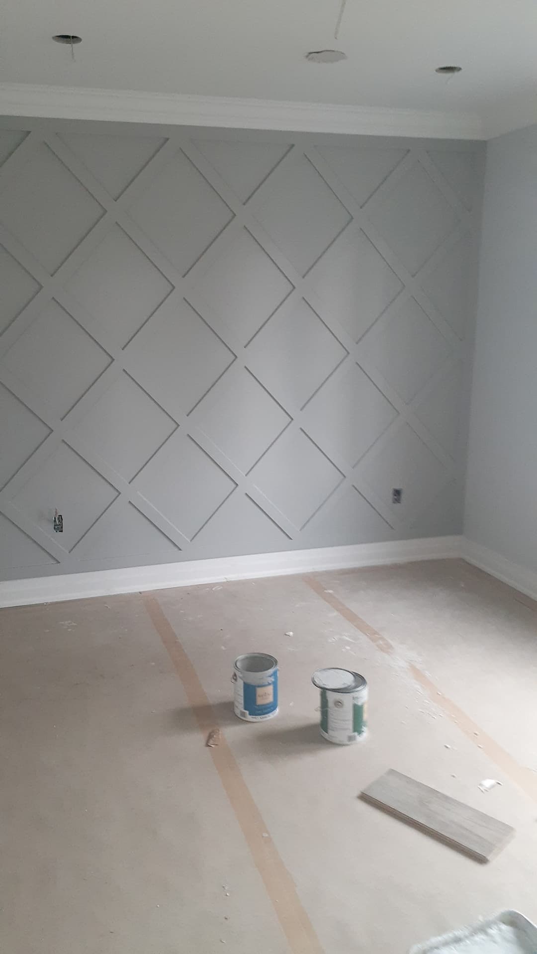 Interior room showing a light gray accent wall with diagonal trim arranged in a repeating diamond lattice pattern. Paint cans and renovation materials sit on the protected floor.