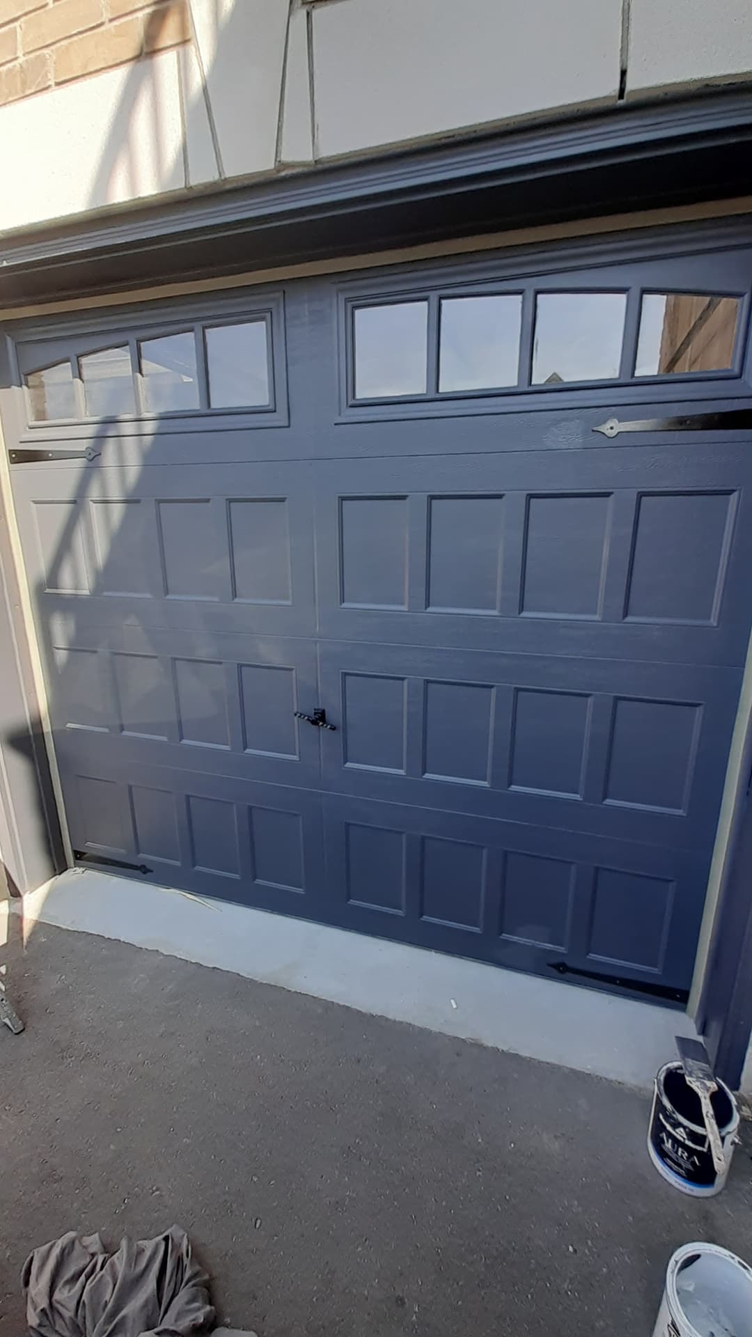 Exterior close-up of a dark blue garage door with rectangular panel detailing and a row of small upper windows. Painting supplies are visible near the bottom edge.