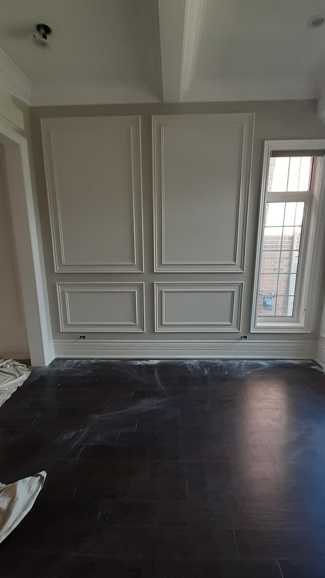 Interior room with a formal wall treatment made of large rectangular molding panels painted a soft gray-beige. A tall window is on the right, and dark hardwood flooring is covered with renovation dust.