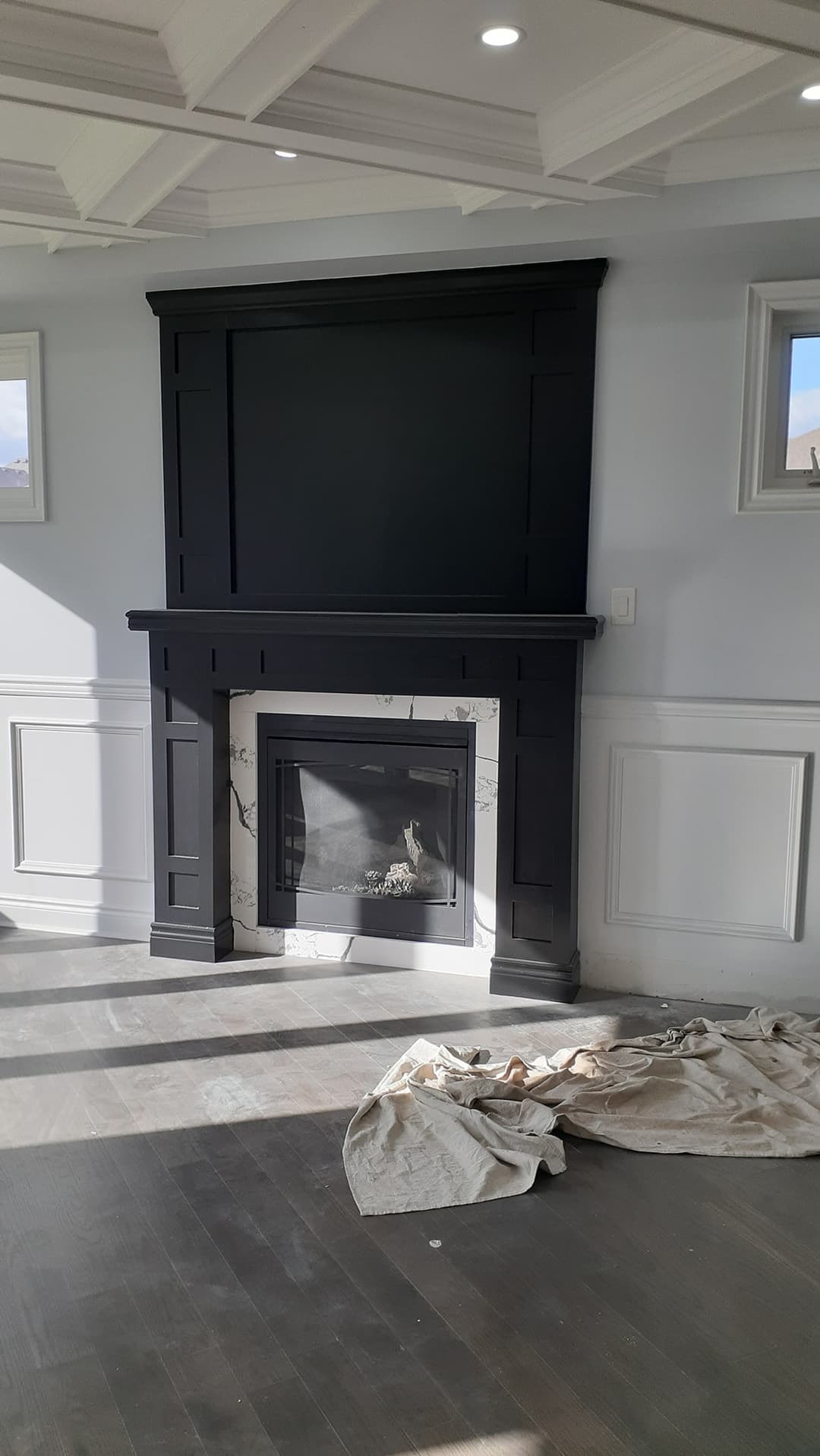 Living room with a black paneled fireplace surround and mantel, white marble-style fireplace insert, light gray walls, white wainscoting, coffered ceiling, recessed lighting, and dark hardwood floors.