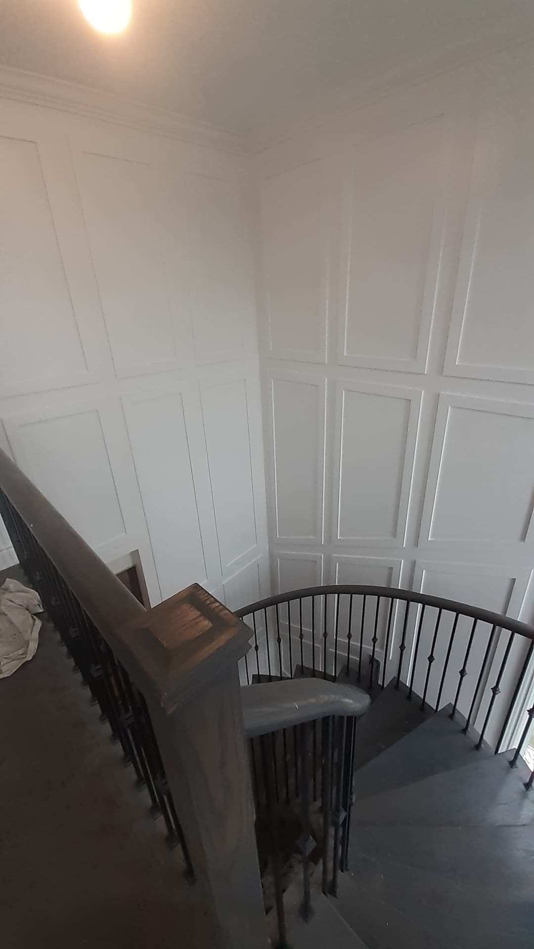 Stairwell with tall white paneled wall molding extending down the staircase. A curved black metal railing and dark wood handrail follow the stairs.