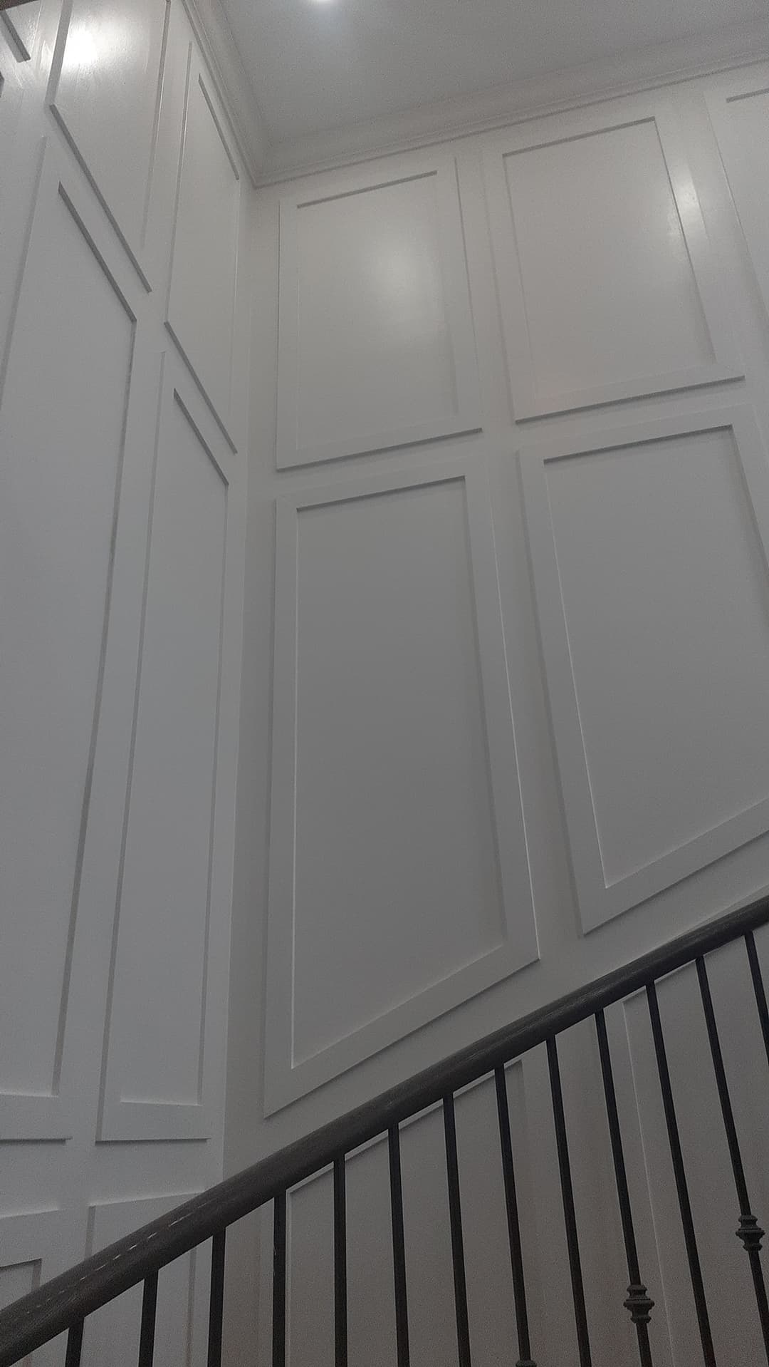 Close-up view of a tall staircase wall with large white rectangular panel molding. A dark handrail and black metal balusters run diagonally across the lower part of the image.